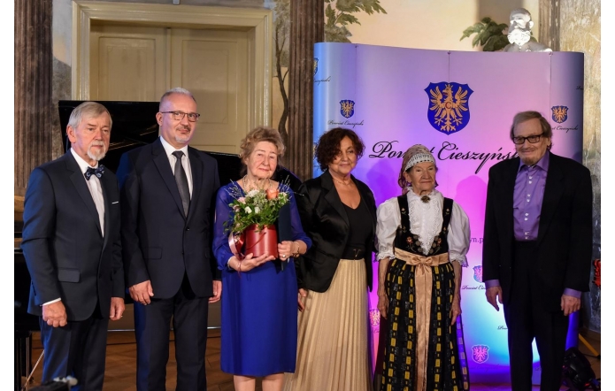Sześć osób stojących w szeregu. Od lewej: od lewej Stanisław Kubicius, Mieczysław Szczurek, prof. Heska-Kwaśniewicz, Janina Żagan, Helena Wróbel, Adam Makowicz. Dwie kobiety znajdują się po środku, jedna z nich trzyma w rękach czerwony wazon z kwiatami. Kobieta po prawej stronie ubrana jest w tradycyjny strój ludowy. Pozostałe osoby mają na sobie eleganckie stroje, głównie garnitury. W tle znajduje się niebieska ścianka z herbem oraz napisem "Powiat Cieszyński".