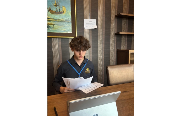 Chłopak z identyfikatorem na niebieskiej smyczy siedzi przy stole i przegląda dokumenty. Przed nim stoi laptop, a w tle widoczna jest kartka z informacją o stażu uczniowskim oraz obraz na ścianie.