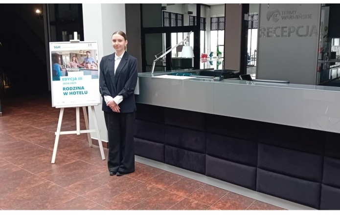 Ewa Raszka w eleganckim stroju hotelowym stoi przy recepcji Term Warmińskich. Obok znajduje się plakat promujący program "Rodzina w hotelu" organizowany przez SGH. Scena oddaje atmosferę profesjonalizmu i gościnności.