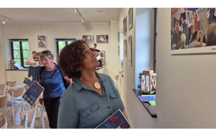 Janina Żagan ogląda z zainteresowaniem wystawę fotograficzną o folklorze. W dłoni trzyma program wydarzenia. W tle kobieta gra na skrzypcach, a inna uczestniczka przegląda materiały. Wnętrze jasne, pełne regionalnych akcentów.