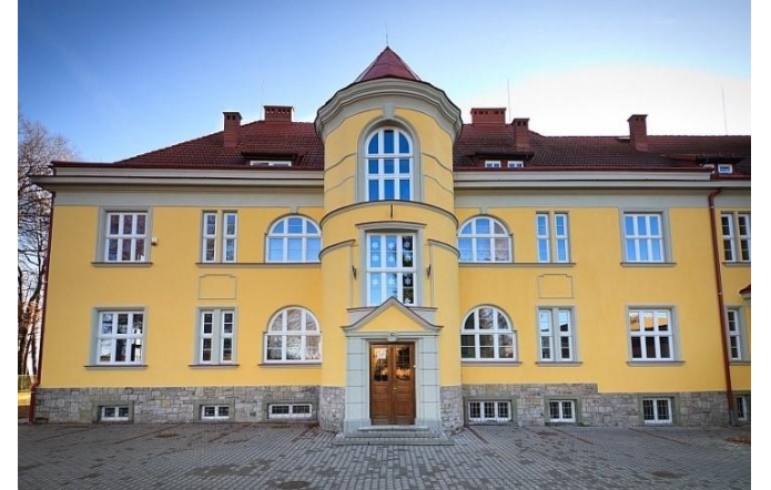 Symetryczny, dwukondygnacyjny Budynek główny Zespołu Szkół Przyrodniczo-Technicznych w Międzyświeciu, o żółtej elewacji z jasnymi obramowaniami okien, czerwonym dachem i centralną, lekko wysuniętą wieżą z półokrągłym zwieńczeniem; przed wejściem niewielki plac z kostki brukowej. 