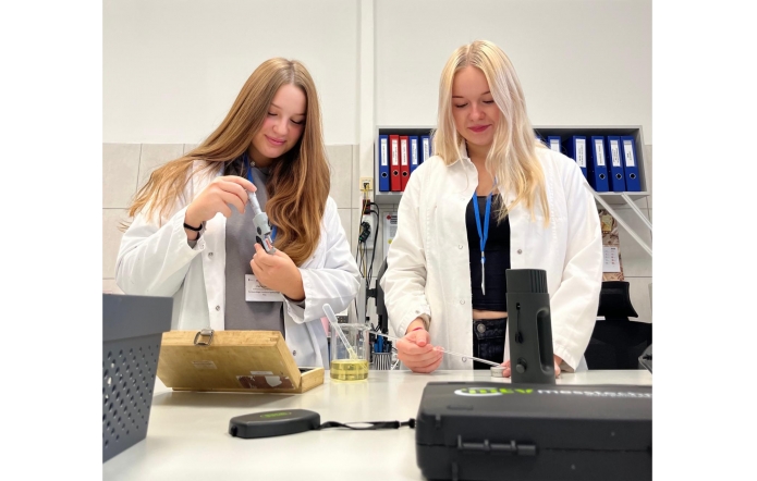 Dwie uczennice w fartuchach laboratoryjnych pracują przy stole w pracowni. Jedna korzysta z mikrometru do pomiaru elementu, druga obsługuje sprzęt pomiarowy przy użyciu sondy. Na blacie znajdują się naczynia laboratoryjne z cieczą oraz aparatura pomiarowa, a w tle widać segregatory z dokumentacją.