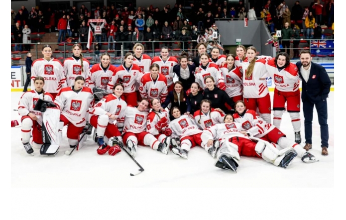 Grupa młodych kobiet w sportowych strojach z biało-czerwonymi akcentami stoi i klęczy na lodowisku. Na ich koszulkach widnieje godło Polski. Są uśmiechnięte i zadowolone, otoczone przez dwie osoby w eleganckich strojach oraz trenera. W tle widać trybuny z kibicami trzymającymi flagi i transparenty. Atmosfera wyraża radość i dumę z osiągnięcia sportowego sukcesu.