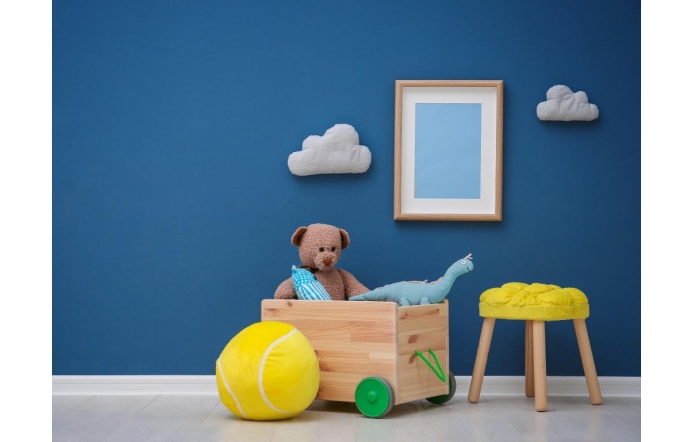 Pokój dziecięcy z niebieską ścianą, ozdobiony białymi chmurkami i ramką. Na podłodze drewniany wózek z pluszowym misiem i dinozaurem, obok duża żółta piłka. Po prawej stoi mały taboret z żółtym siedziskiem.