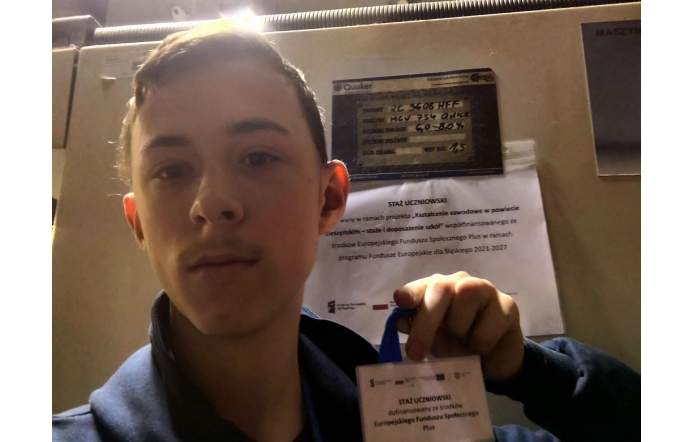 Chłopak robi zdjęcie typu selfie i pokazuje identyfikator z napisem „Staż uczniowski”. W tle widoczna jest maszyna oraz kartka informacyjna dotycząca projektu.