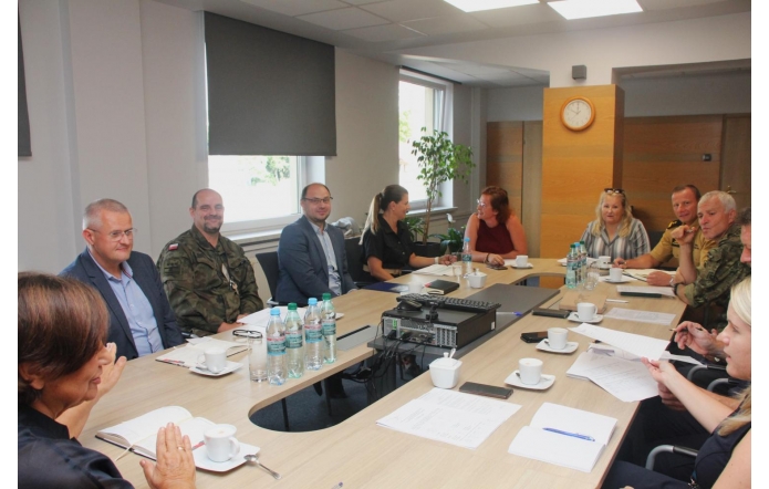 Przy dużym stole w sali konferencyjnej siedzi grupa osób – część w mundurach wojskowych, część w ubraniach cywilnych. Na stole stoją butelki wody, filiżanki z kawą i notatniki, a w centralnym miejscu leży sprzęt multimedialny