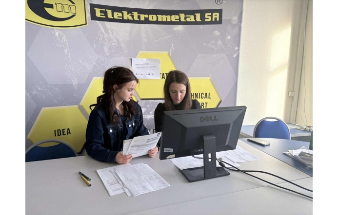 Dwie młode dziewczyny siedzą przy biurku i pracują przy komputerze. Na blacie znajdują się dokumenty oraz telefon, a nad zdjęciem widoczny jest żółty pasek z nazwą firmy Elektometal.