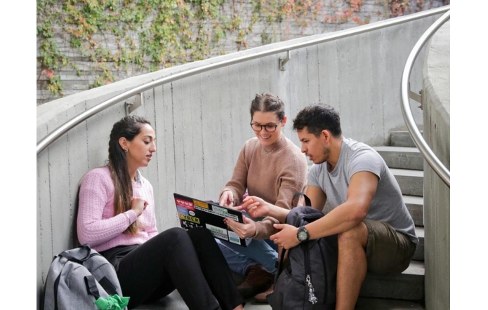 Troje młodych ludzi siedzi na betonowych schodach w otwartym, zielonym otoczeniu. Mają przy sobie plecaki, a jedna z osób trzyma laptop, nad którym wspólnie dyskutują i wskazują coś na ekranie.