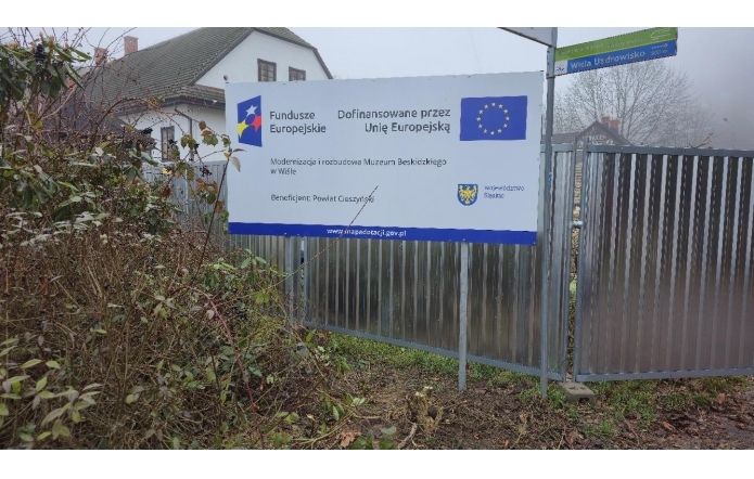 Metalowe ogrodzenie z zamontowaną tablicą informacyjną o dofinansowaniu z Unii Europejskiej. W tle budynek muzeum, krzewy i drzewa bez liści. Po prawej stronie widoczny fragment bramy i znak drogowy.