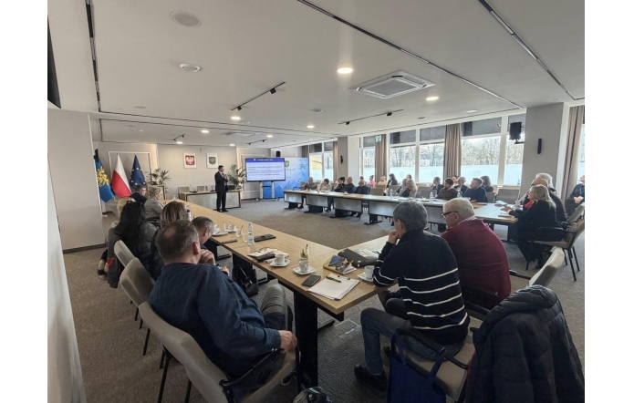 W sali sesyjnej kilkanaście osób siedzi przy dużym stole ustawionym w kształt litery U. Z przodu stoi mężczyzna obok ekranu z wyświetlonym slajdem, a w tle widoczne są flagi i godła. Na stołach znajdują się filiżanki, butelki wody, notatniki i telefony.