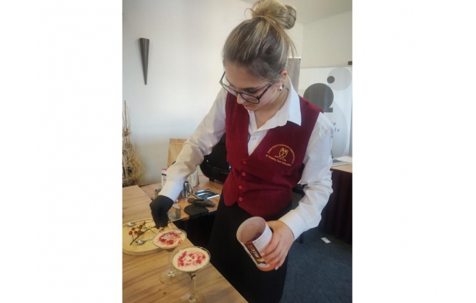 Młoda dziewczyna w eleganckim stroju kelnerskim, z bordowym gorsetem i białą koszulą, przygotowuje dwa koktajle ozdobione różowym proszkiem. Ma związanego w kok włosy, nosi okulary i czarną rękawiczkę na jednej dłoni, pracując przy drewnianym blacie.