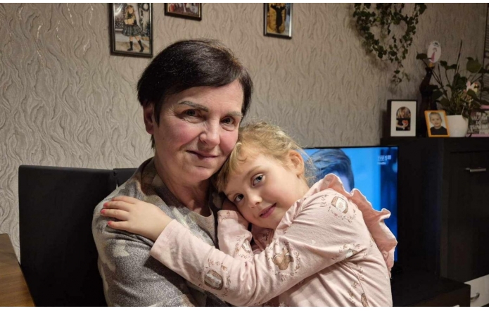 Starsza kobieta z krótkimi, ciemnymi włosami i łagodnym uśmiechem trzyma w ramionach małą dziewczynkę z jasnymi włosami. Dziewczynka, ubrana w różową bluzkę, przytula się z miłością do kobiety, spoglądając spokojnie przed siebie. W tle widać wnętrze mieszkania, na ścianie znajdują się zdjęcia oraz rośliny ozdobne. Atmosfera zdjęcia jest ciepła i rodzinna.
