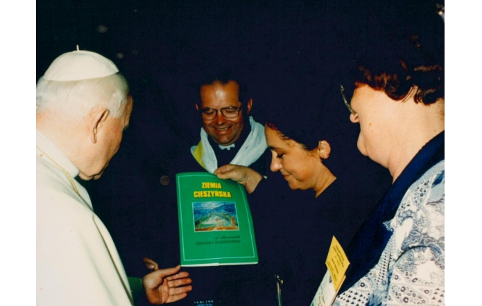 Janina Żagan wręcza papieżowi Janowi Pawłowi II książkę pt. „Ziemia Cieszyńska”. Obok stoją duchowny i kobieta z identyfikatorem. Wszyscy uśmiechnięci, skupieni na symbolicznym geście.