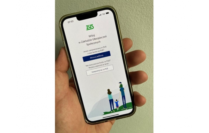 Telefon trzymany w lewej dłoni wyświetla ekran powitalny aplikacji ZUS (Zakład Ubezpieczeń Społecznych). Na górze znajduje się logo ZUS i tekst powitalny. Poniżej widoczne są dwa przyciski: „Aktywuj aplikację” oraz „Zarejestruj się na PUE”. W dolnej części ekranu widać rysunek rodziny – dwoje dorosłych i dwójka dzieci.