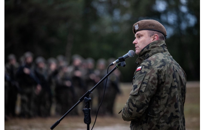 Żołnierz w mundurze polowym mówi do mikrofonu podczas zbiórki, w tle widoczna grupa wojskowych ustawionych w szeregu na otwartym terenie.