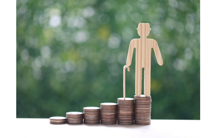 Drewniana figurka starszej osoby z laską stoi na najwyższym z kilku stosów monet ustawionych obok siebie w rosnącym układzie, na tle rozmytej zieleni.