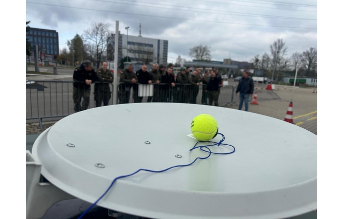 ChatGPT powiedział:  Żółta piłka tenisowa z przywiązanym niebieskim sznurkiem leży na białej antenie satelitarnej. W tle grupa umundurowanych osób stoi za barierką na placu, obok widoczne są pachołki drogowe i biurowce.