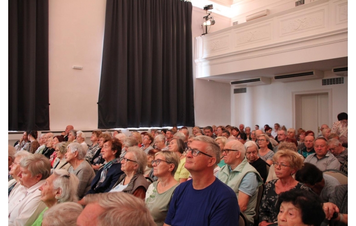 Sala pełna osób starszych, siedzących blisko siebie na krzesłach, uważnie patrzących w stronę sceny lub ekranu. Wnętrze jasne, z wysokim sufitem, dużymi zasłonami i balkonem u góry.