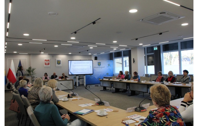 Nowoczesna sala sesyjna z dużymi oknami i jasnym oświetleniem. Przy stołach w kształcie litery „U” siedzą uczestnicy spotkania, słuchając gościa przy mównicy z lewej strony. Na środku stoi ekran z prezentacją, a w tle widoczne są flagi Polski, Unii Europejskiej i województwa śląskiego oraz herby na ścianie.