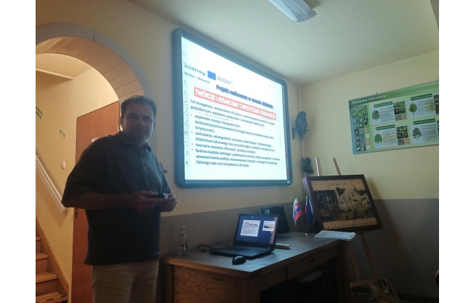 Osoba stoi po lewej przy łukowym przejściu. Na ścianie świeci ekran z prezentacją z ikonami i strzałkami. Przed nim biurko z laptopem i sprzętem; na blacie małe flagi. Na ścianie obok wisi mapa/obraz.