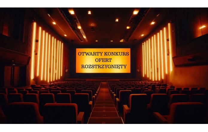 Duża, pusta sala kinowa z długimi rzędami foteli ustawionych amfiteatralnie, skierowanych w stronę szerokiego ekranu. Na ekranie wyświetlony jest napis „OTWARTY KONKURS OFERT ROZSTRZYGNIĘTY”. Wnętrze wypełnia ciepłe, pomarańczowe światło, które podkreśla spokojny i oficjalny charakter miejsca.