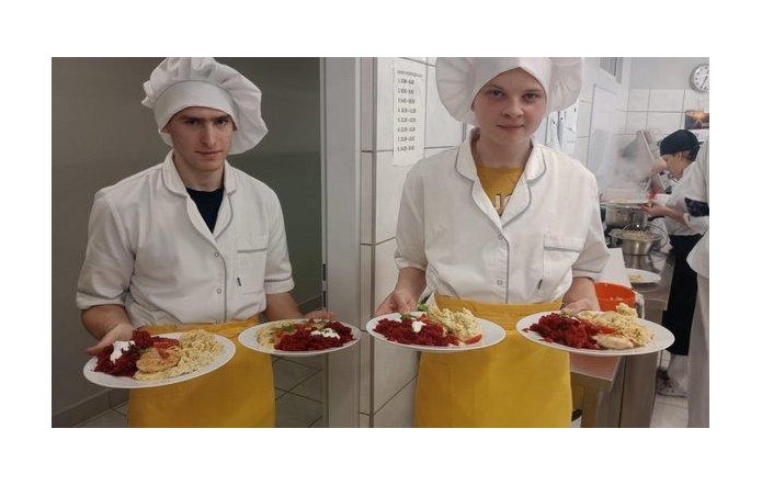 Dwoje młodych kucharzy w białych fartuchach i czapkach kucharskich trzyma po dwa talerze z kolorowymi daniami. W tle widać kuchnię szkolną, uczniów przy pracy i parujący garnek na kuchence.