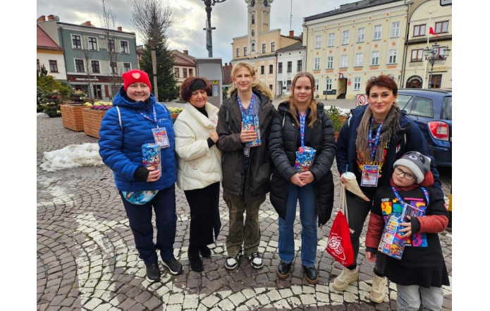 Grupa sześciu osób, w różnym wieku, druga od lewej – Janina Żagan, Starosta, stoi na brukowanym placu w centrum miasta. W rękach trzymają kolorowe puszki z logo Wielkiej Orkiestry Świątecznej Pomocy, uśmiechają się i mają przypięte identyfikatory. W tle widać kamienice, ratusz i dekoracje zimowe. Atmosfera pełna zaangażowania i życzliwości.
