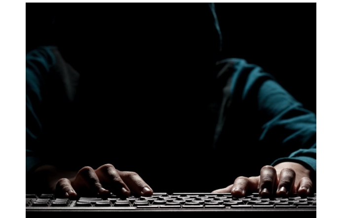 Osoba w kapturze siedzi przy komputerze w ciemnym otoczeniu, a oświetlenie podkreśla jedynie dłonie piszące na klawiaturze.