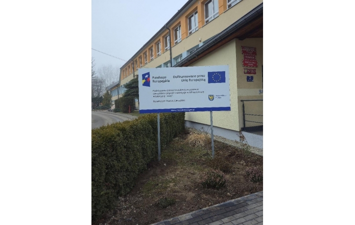 Poziomy baner informacyjny ustawiony na metalowych słupkach przed budynkiem użyteczności publicznej. Na banerze umieszczone są logotypy Funduszy Europejskich, Unii Europejskiej oraz Województwa Śląskiego. Widoczny jest napis „Dofinansowane przez Unię Europejską” oraz informacja o realizacji projektu. W tle znajduje się elewacja budynku z oknami, chodnik oraz teren zielony.