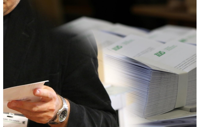 Osoba w ciemnym stroju trzyma w dłoni białą kartkę papieru, skupiając się na jej zawartości. W tle widoczny jest stos ułożonych równo dokumentów z zielonym logo, sugerujących oficjalny charakter. 