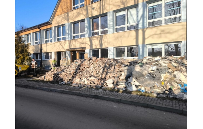 Przed dwukondygnacyjnym budynkiem z jasną elewacją i rzędami okien znajduje się dużo gruzu budowlanego z cegieł, fragmentów tynku, płyt oraz elementów sanitarnych. Przy wejściu do budynku stoją dwie osoby, jedna w odzieży roboczej. Po lewej stronie widoczna jest niewielka maszyna budowlana. Wzdłuż gruzu przebiega chodnik i jezdnia.