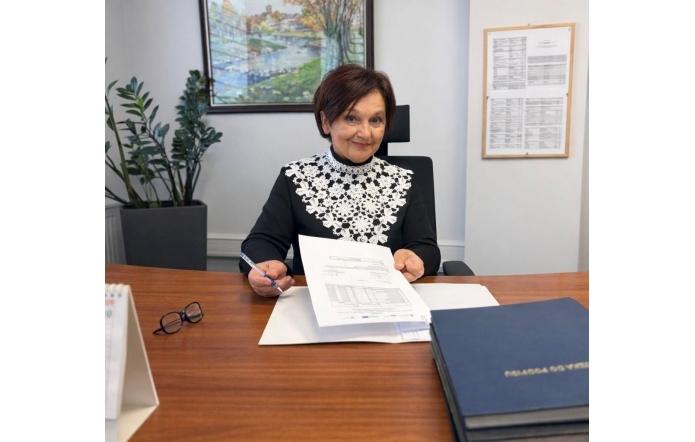 Janina Żagan Starosta Cieszyńska siedzi przy biurku w gabinecie i trzyma w dłoni dokument. Na biurku leżą segregatory, okulary oraz kalendarz, a w tle widać rośliny doniczkowe i obraz na ścianie.