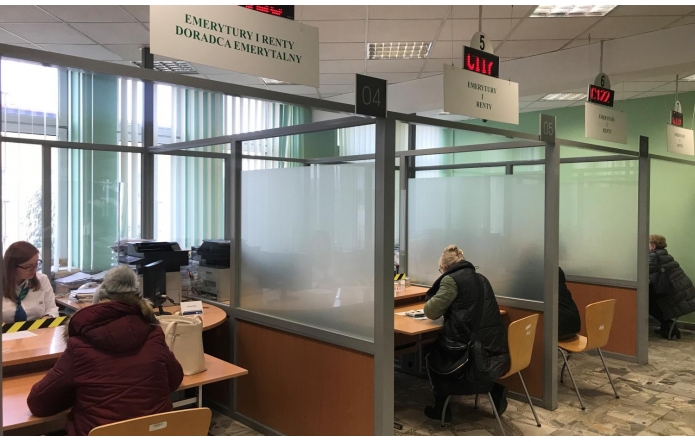 Wnętrze urzędu z boksami obsługi, gdzie klienci siedzą przy biurkach i załatwiają sprawy z pracownikami, w otoczeniu zielonych ścian, matowych przegród i elektronicznych wyświetlaczy numerków.