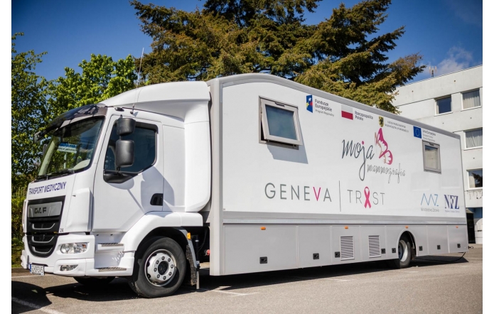 Biały mammobus — mobilna pracownia mammografii — zaparkowany na słonecznym parkingu. Na boku widoczne logotypy,  w tle zielone drzewa, fragment budynku i błękitne niebo.