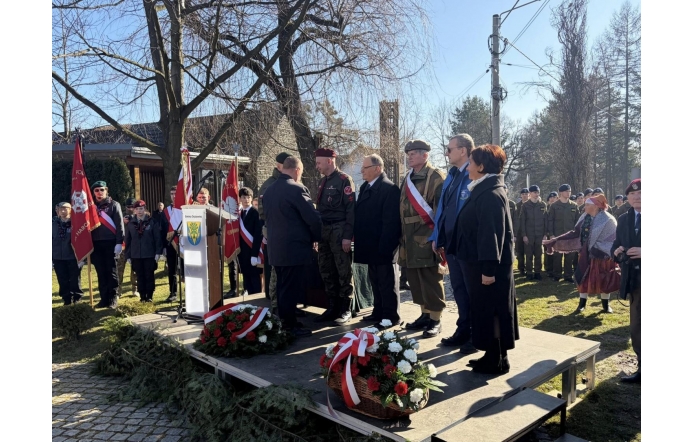 Na niewielkiej scenie stoi grupa osób, w tym mężczyźni w mundurach i garniturach oraz kobieta w płaszczu. Dwóch mężczyzn podaje sobie ręce obok mównicy z herbem gminy. Na scenie leżą wieńce z czerwonych i białych kwiatów, a w tle stoją harcerze, żołnierze i sztandary.