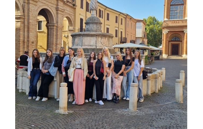 Grupa nastolatek stoi wokół kamiennej, zabytkowej fontanny na brukowanym placu. Wokół arkady z cegły, jasne kamienice, parasole kawiarniane i fasada kościoła.