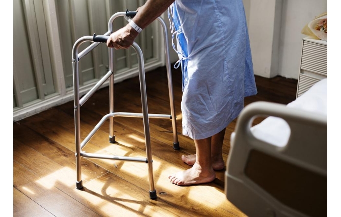 Starsza osoba w szpitalnym fartuchu stoi boso przy łóżku, opierając się na metalowym chodziku. Widać drewnianą podłogę i fragment szpitalnego łóżka, wokół panuje spokojna, poranna cisza.