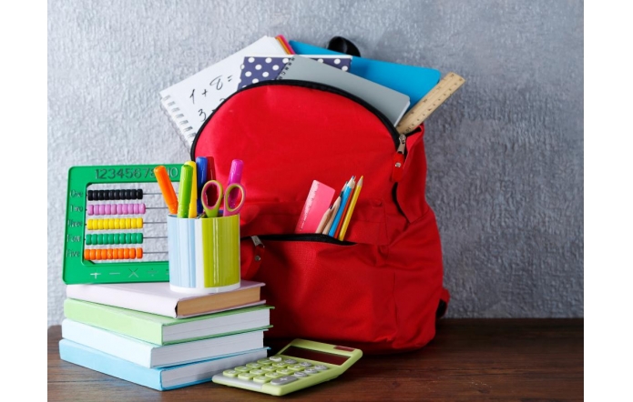 Czerwony plecak wypchany zeszytami, książkami i przyborami szkolnymi stoi na drewnianym stole obok stosu kolorowych książek, kubka z długopisami, liczydła i kalkulatora.