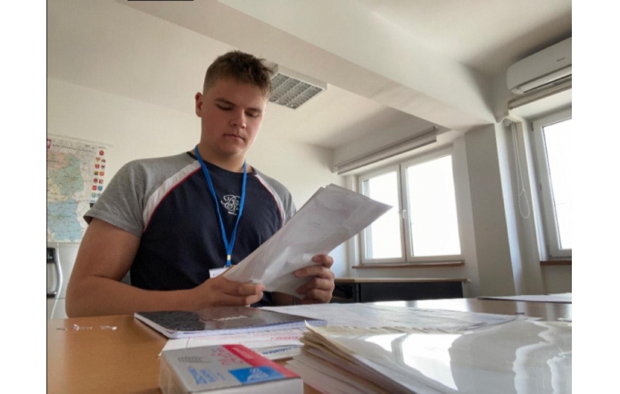 Młody mężczyzna w koszulce i z identyfikatorem na szyi stoi przy biurku w jasnym biurze. Trzyma w rękach dokumenty, nad którymi się pochyla, wokół widać papiery, mapę i biurowe wyposażenie.