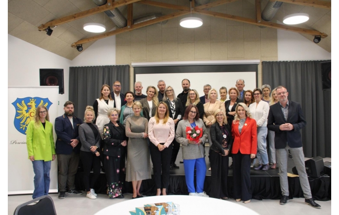 Grupowe zdjęcie uczestników spotkania, ustawionych na scenie w dwóch rzędach; większość uśmiechnięta, w centrum kobieta trzyma bukiet kwiatów. W tle ekran i zasłony sceniczne, po lewej roll-up z herbem Powiatu Cieszyńskiego. Nowoczesna sala z widoczną konstrukcją drewnianego stropu.