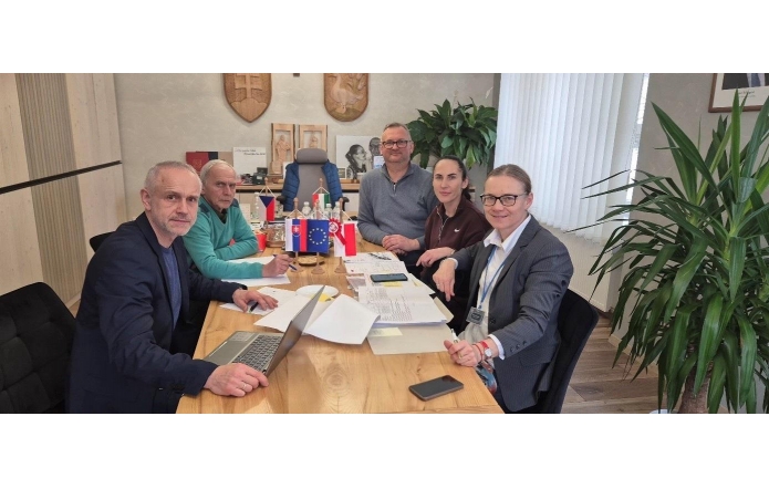 Pięć osób siedzi przy drewnianym stole w biurze. Na stole leżą dokumenty, laptop, telefon i małe flagi państwowe oraz Unii Europejskiej. W tle widoczne są rośliny doniczkowe, okno z żaluzjami oraz elementy wystroju biura.