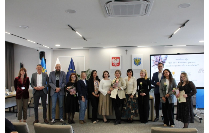 Grupa osób stojących w sali sesyjnej i pozujących do wspólnego zdjęcia. Część z nich trzyma kwiaty i dyplomy, a w tle widoczne są flagi, herby oraz ekran z prezentacją.