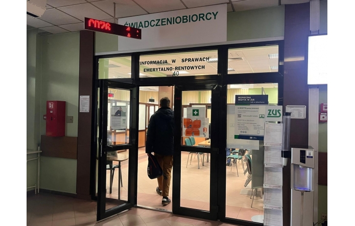 Wejście do sali obsługi ZUS z napisem „ŚWIADCZENIOBIORCY” i „Informacja w sprawach emerytalno-rentowych”. Mężczyzna w ciemnej kurtce wchodzi przez szklane drzwi, trzyma torbę. W środku widać krzesła, okienka, elektroniczny wyświetlacz kolejkowy oraz plakaty informacyjne na szybach.