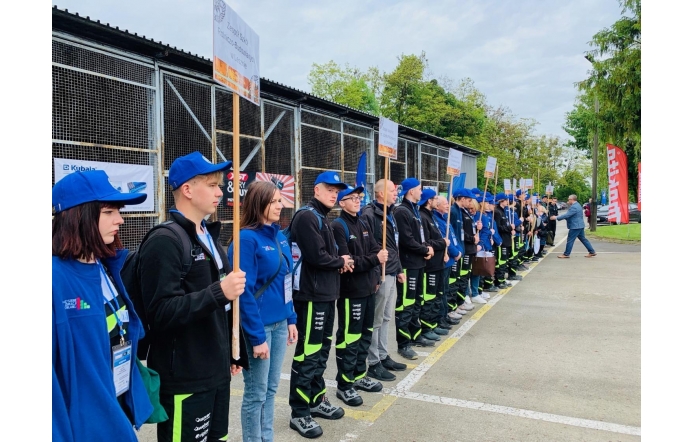 Grupa młodych osób w jednakowych strojach i niebieskich czapkach stoi w szeregu na zewnątrz, trzymając tabliczki z nazwami szkół. Uczestniczą w Olimpiadzie „Buduj z Pasją”.