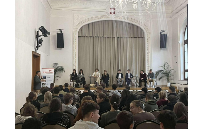 W przestronnej, jasnej sali z wysokim sufitem i żyrandolem na scenie siedzi kilka osób ustawionych w szeregu na krzesłach. Przed nimi zgromadzona jest duża grupa młodych ludzi. Po lewej stronie stoi mężczyzna przemawiający obok roll-upu, a po bokach sceny ustawiono rośliny w donicach.