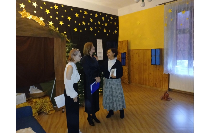 Trzy kobiety rozmawiają (pierwsza od prawej Janina Żagan, Starosta Cieszyński) w pomieszczeniu o ciepłej, przytulnej atmosferze. Tło zdobi ciemna dekoracja z motywem nocnego nieba, pełna żółtych gwiazd i świetlistej komety. Obok stoi choinka, a ściany z drewnianymi panelami i żółtym kolorem tworzą ciepły klimat. Na podłodze widoczne są rekwizyty, które nadają przestrzeni teatralnego charakteru.