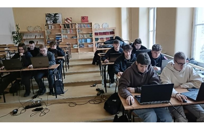 Grupa uczniów siedzi w klasie przy drewnianych ławkach, pracując na laptopach. W tle regały z książkami i dekoracje ścienne. Jasne ściany i duże okna wpuszczają naturalne światło. Niektórzy uczniowie w bluzach z kapturem, skupieni na ekranach komputerów.