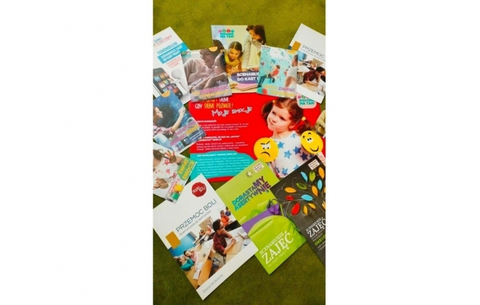 Kolorowe broszury edukacyjne rozłożone na zielonym tle przedstawiają materiały poświęcone emocjom, przemocy, asertywności, fonoholizmowi i rodzinie. Część z nich zawiera scenariusze zajęć, ilustracje dzieci i edukacyjne grafiki.