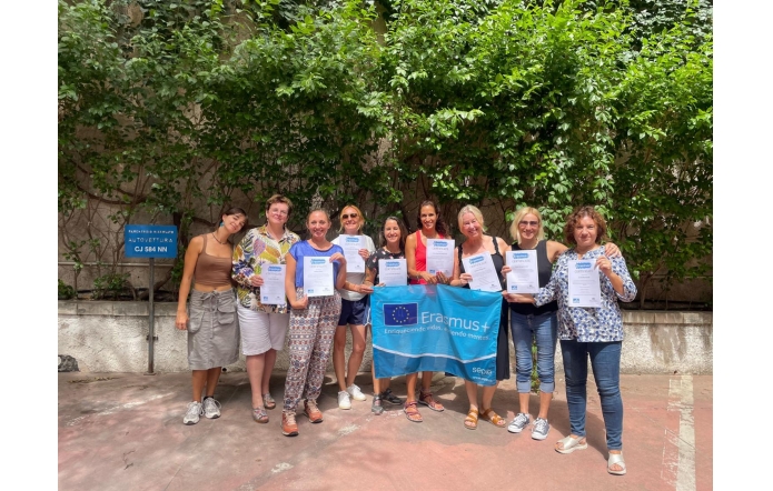 Dziesięć uśmiechniętych kobiet pozuje na dworze z certyfikatami, a pośrodku trzymają niebieską flagę Erasmus+. Za grupą zielona ściana pnączy, słoneczna, radosna atmosfera.