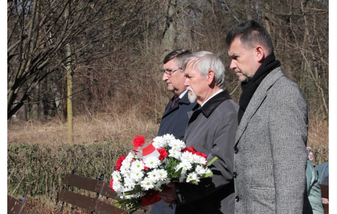 Trzech mężczyzn w eleganckich płaszczach i szalikach stoi w skupieniu – od prawej: Jacek Wiśniewski, sekretarz Powiatu, Stanisław Kubicius, Przewodniczący Rady Powiatu i Jerzy Pilch, członek Zarządu Powiatu. Stanisław Kubicius trzyma biało-czerwony wieniec z kwiatami, przewiązany wstążką w barwach narodowych. W tle ławki, przycięte krzewy i bezlistne drzewa, sugerujące chłodną porę roku.